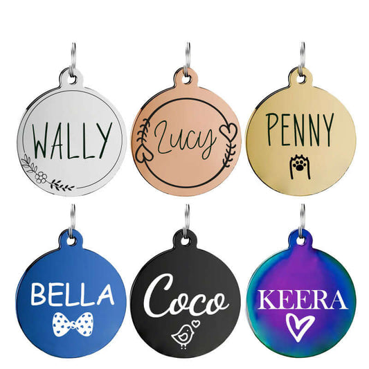 Assorted round stainless steel dog tags in silver, gold, rose gold, black, blue, and rainbow finishes engraved with names like Wally, Lucy, Penny, Bella, Coco, and Keera, featuring unique icons like floral, heart, bow tie, paw print, and bird designs.